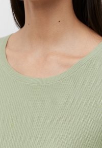 Lichtgroen wafelgebreid topje met een brede halslijn en textuurstof. Zichtbare stiksels langs de halslijn en de randen van de zoom.