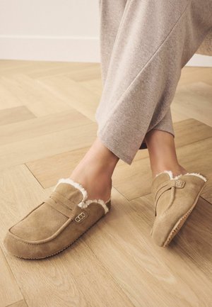 Tan ruskindsindslipper med en blød hvid pelsforing, lukket hæl-design og dekorative stropforstærkninger. Uforet med en struktureret, skridsikker sål.