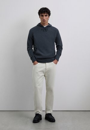 Young man standing with hands in pockets, wearing a dark gray hoodie, off-white pants, and black lace-up shoes, against a plain background.