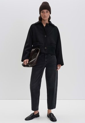 Black cropped jacket with a collar, two front pockets, and button details, paired with dark wash jeans and a brown beanie. Holding a brown bag.