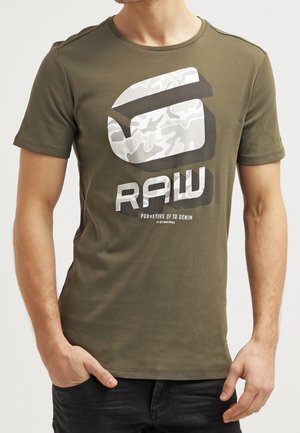 Hombre con camiseta verde oliva con un gran logo de camuflaje blanco y gris y texto "RAW Purveyors of 3D Denim" en el pecho.