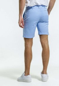 Shorts en coton bleu clair avec une taille plate, un ourlet retroussé et une poche arrière. Associés à des baskets blanches. Poils de jambe et texture de peau visibles.
