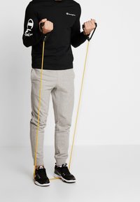 Schwarzes Langarmshirt mit weißem Logo, graue Jogginghose und schwarze Sneakers, in jeder Hand gelbe Widerstandsbänder haltend.