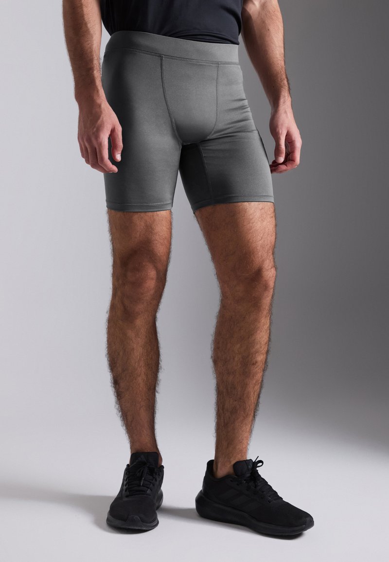 Grå treningsshorts med tettsittende passform, glatt stoff, elastisk midjebånd og flate sømmer. Brukt med svarte treningssko.