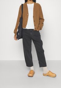 Cárdigan de punto marrón con botones en la parte delantera, camiseta blanca, jeans negros de pierna ancha y zuecos color marrón claro con agujeros; combinado con calcetines grises y una bolsa tote negra.