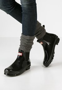 Czarne gumowe botki za kostkę z błyszczącym wykończeniem. Posiadają elastyczne panele boczne i charakterystyczną etykietę "HUNTER" w czerwono-białych kolorach. Noszone z szarymi ocieplaczami na nogi.