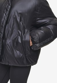 Zwarte pufferjack met een glanzende afwerking, oversized pasvorm, drukknoopsluiting en een ritszak. Logo in contrasterende tekst op de mouw.