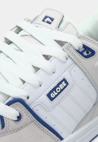 Gros plan sur une basket Globe blanche avec des accents bleus et des lacets blancs, mettant en valeur le logo de la marque et les détails de la couture.