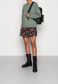 Felpa verde chiaro con polsini a costine, skort nero con motivo floreale e stivali da combattimento neri con lacci. La modella tiene uno zaino nero.