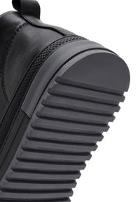 Semelle en caoutchouc noire avec des rainures horizontales profondes et un bord texturé sur une chaussure en cuir noir, inclinée vers le haut sur un fond blanc.
