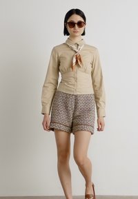 Light beige button-up blouse with a tied neck scarf; patterned brown shorts with contrasting trim, and oversized sunglasses.