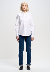 Camisa blanca de botones con cuello, mangas largas y bajo curvado, combinada con jeans ajustados de color azul oscuro y zapatos negros sin cordones.