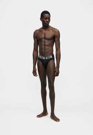 Μυώδης άνδρας με σκούρο δέρμα στέκεται ξυπόλητος φορώντας μαύρα εσώρουχα Calvin Klein, κοιτάζοντας ελαφρώς δεξιά σε απλό φωτεινό φόντο.