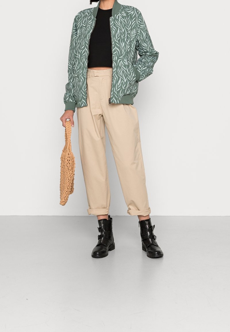 Green bomber jacket with white leaf pattern, black fitted top, beige wide trousers, and black ankle boots with buckle straps. Holding a woven bag.
