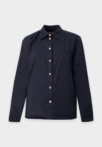 Chemise bleu marine manches longues à boutons avec boutons décoratifs dorés et col classique pointu sur un fond uni.