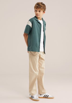 Dječak u bež hlačama, bijelim tenisicama, bijeloj majici i zelenoj košulji kratkih rukava s kopčanjem gleda ustranu s rukama na leđima.