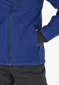 Blaue Outdoor-Jacke mit glatter Textur, Reißverschluss vorne und seitlichen Taschen. Verfügt über schwarze Akzente an den Bündchen zur Anpassung.