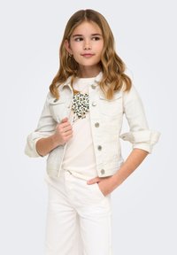 Κορίτσι με μακριά κυματιστά μαλλιά που φοράει λευκό μπουφάν, λευκό παντελόνι και πουκάμισο με πολύχρωμη καρδιά, ποζάροντας με το ένα χέρι στην τσέπη.