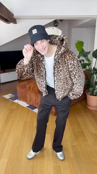 Leopardikuosinen turkistakki vetoketjulla, valkoinen t-paita, mustat housut ja hopeiset urheilukengät. Musta lippalakki logolla. Sisätilassa.