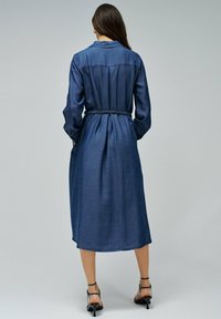 Vestido midi de mezclilla con mangas largas, cintura ceñida y corte holgado. La tela es de color azul oscuro, con una textura suave y detalles de botones.