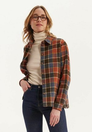 Veste en flanelle à carreaux en laine dans des tons de marron, bleu et rouille, avec une fermeture à boutons, un col et une coupe décontractée. Portée par-dessus un col roulé beige.