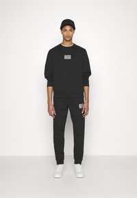 Felpa nera in cotone e pantaloni jogger abbinati, dotati di toppe con logo, vestibilità comoda, polsini a coste, e completati da sneaker bianche.