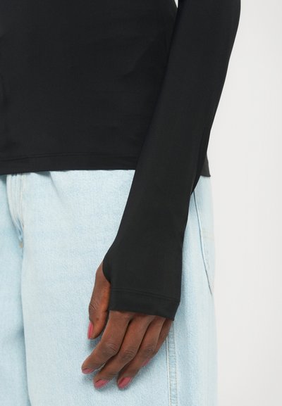 Maglietta nera a maniche lunghe aderente con una texture liscia, dotata di fori per il pollice nei polsini, abbinata a jeans in denim azzurro chiaro.