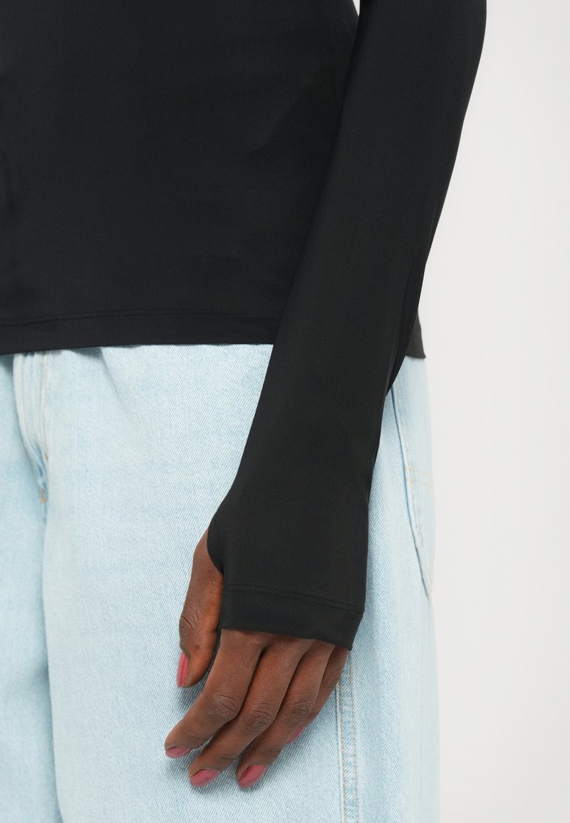 Haut noir ajusté à manches longues avec une texture lisse, présentant des trous pour les pouces aux poignets, associé à un jean en denim bleu clair.