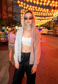 Felpa grigia con zip sopra una maglietta crop bianca, abbinata a pantaloni jogger in satin neri; sullo sfondo ci sono lanterne arancioni illuminate di notte.