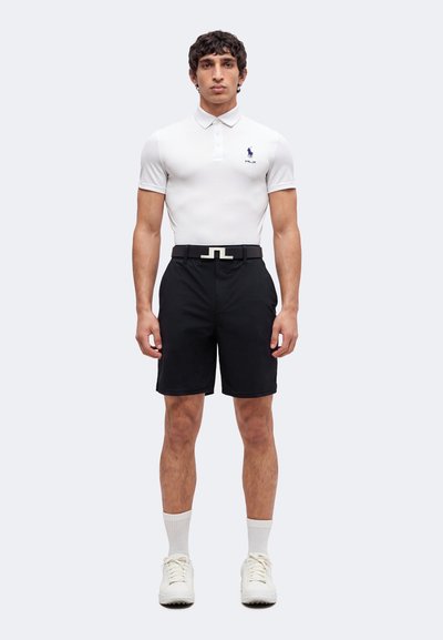 Homme debout portant un polo blanc, un short noir avec ceinture, des chaussettes blanches et des baskets blanches sur un fond clair uni.