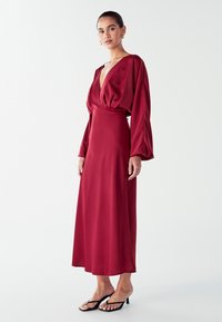 Long burgundy satin dress with a deep V-neck and balloon sleeves, featuring a fitted waist and flowy skirt. Worn with black sandals.