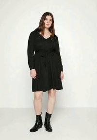 Mulher com cabelo castanho longo, vestindo um vestido preto de manga longa até o joelho com um cinto na cintura, combinado com botas pretas de tornozelo, de pé contra um fundo neutro.