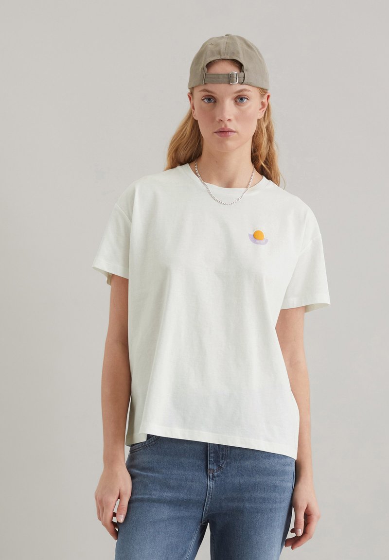 Tricou din bumbac de culoare verde deschis, cu o croială lejeră. Prezintă un mic design brodat cu un ou galben și purpuriu pe partea stângă a pieptului. Blugi vizibili.