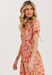 Kvetinové šaty s paletou farieb v ružovej, oranžovej a červenej, s krátkymi rukávmi a priliehavým živôtikom, ktorý prechádza do nariasenej sukne.