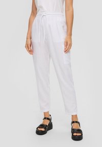 Weiße Leinenhose mit elastischem Bund und Kordelzug, ausgestattet mit Seitentaschen; kombiniert mit schwarzen Plateausandalen. Glatte Textur, lockere Passform.