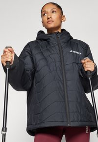 Veste isolante noire avec capuche, présentant un design matelassé et une fermeture éclair. La personne tient des bâtons de randonnée, avec un branding minimal visible.