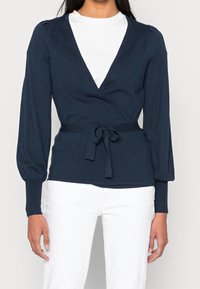 Top avvolgente blu navy con maniche lunghe a sbuffo, nodo in vita e polsini a coste, abbinato a pantaloni bianchi. Tessuto liscio e scollatura a V profonda.