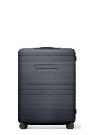 Valise rigide gris foncé avec des rainures horizontales et quatre roues pivotantes noires, présentée fermée et debout sur un fond blanc.