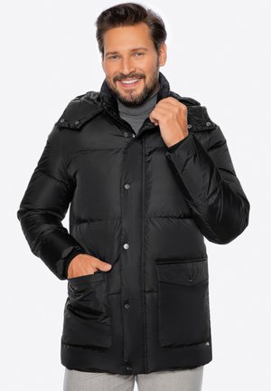 Uomo sorridente che indossa una giacca a piumino nera con bottoni a scatto e cappuccio, mano in tasca, che aggiusta il colletto, su sfondo bianco.