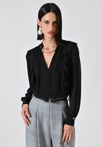 Blusa nera con dettagli arricciati, scollatura a V profonda e maniche lunghe, abbinata a pantaloni a vita alta grigi a quadri e una cintura nera.