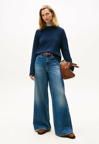 Frau in einem marineblauen Pullover, weiten blauen Jeans, braunen Mokassins und mit einer großen braunen Handtasche vor einem schlichten weißen Hintergrund.