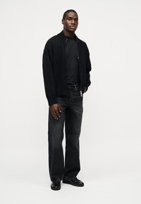 Νέος άντρας που στέκεται, φοράει μαύρο μπουφάν με φερμουάρ, μαύρο μπλουζάκι, σκούρο γκρι τζιν και μαύρα παπούτσια μπροστά σε απλό λευκό φόντο.