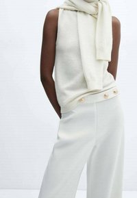 Gehaakte crèmekleurige mouwloze top gecombineerd met hoog waaiervormige lichte broeken met gouden knoopdetails. Zachte textuur met een minimalistisch ontwerp.