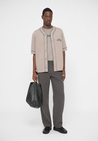 Camicia beige in stile baseball con rifiniture nere, t-shirt grigia, pantaloni grigi e una grande borsa nera in pelle. Il modello indossa sneaker nere.