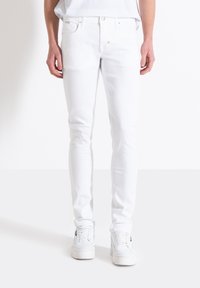 Jeans ajustados blancos de mezclilla, con cinco bolsillos, pasadores para cinturón y cierre de botón en la parte delantera. Llevados con zapatillas blancas.