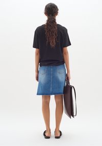 Svart t-shirt med kort ärm, blå denimkjol, platta svarta sandaler, håller en brun toteväska. Rakt, långt hår. Enhetlig vit bakgrund.