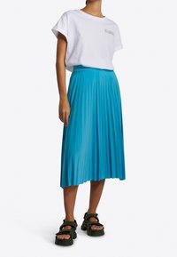Mujer vestida con una camiseta blanca de manga corta metida dentro de una falda midi plisada azul y sandalias negras gruesas, de pie contra un fondo blanco.
