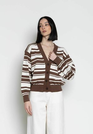 Donna con capelli neri lisci indossa un cardigan a righe marroni e bianche con bottoni e pantaloni bianchi, in piedi su uno sfondo chiaro e uniforme.