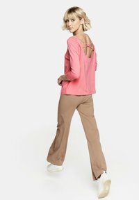 Różowa bluzka z długim rękawem i odkrytymi plecami, z krzyżującymi się paskami, w połączeniu z jasnobrązowymi spodniami dzwonami i białymi trampkami, w pozie do chodzenia.