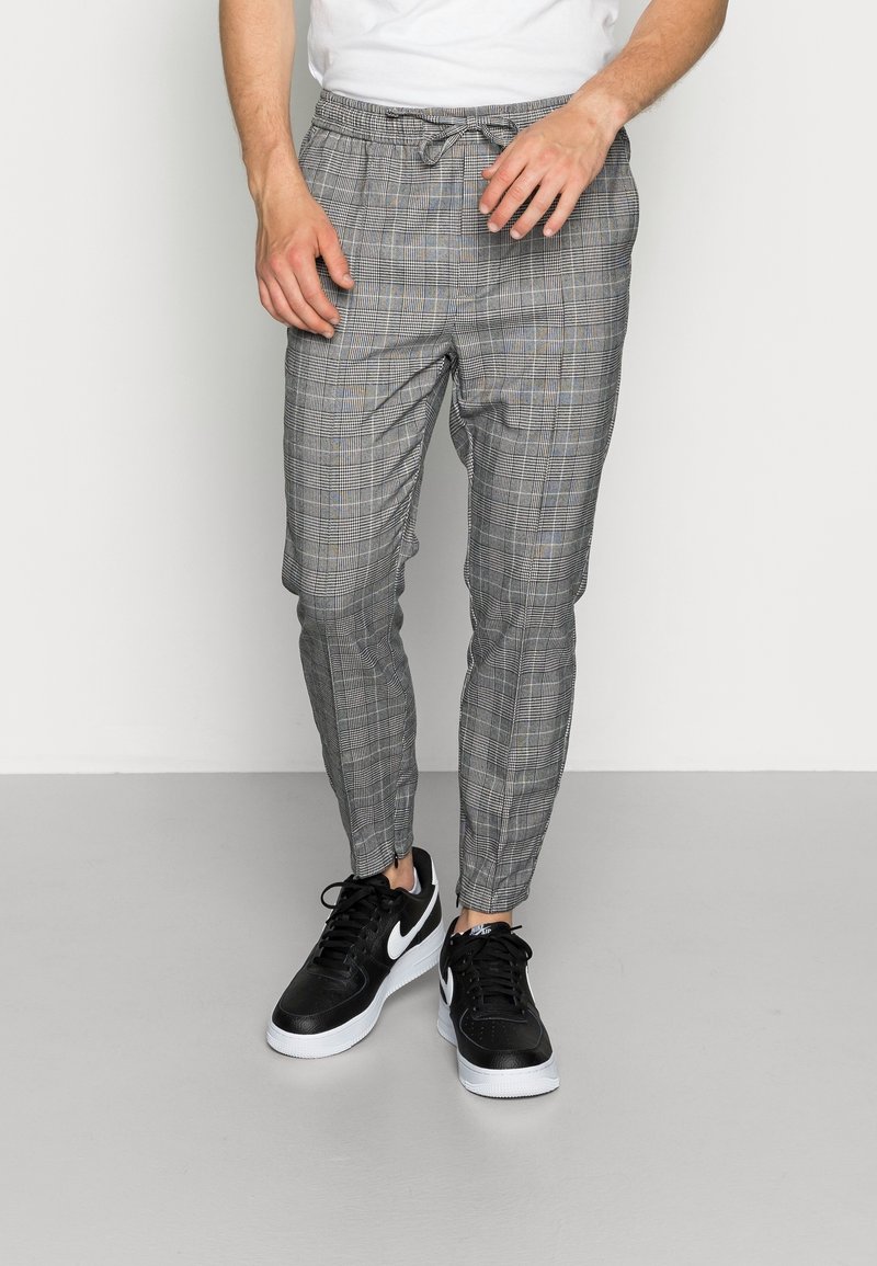 Homme portant un pantalon fuselé à carreaux gris avec une taille à cordon et des baskets Nike noires, debout sur un sol gris contre un mur blanc.